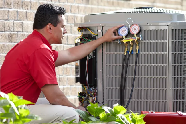 Technician in red shirt checks pressure gauges on outdoor HVAC unit beside brick wall and green plants.