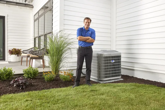Man in blue polo standing outside next to a modern air conditioning unit by a white house with landscaped garden.