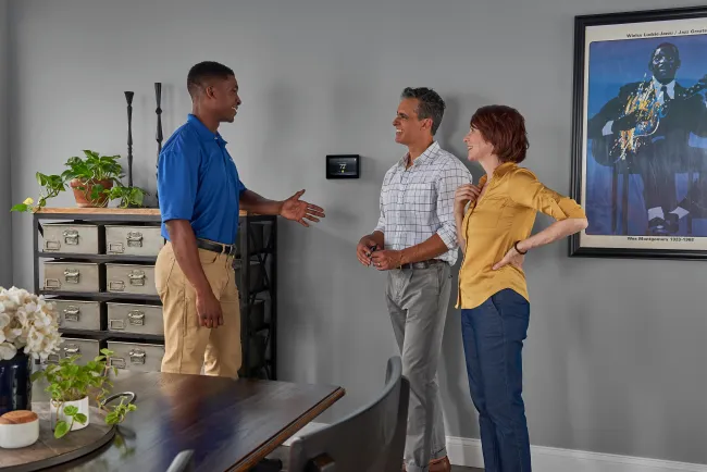 Technician discussing smart thermostat installation with smiling couple in modern home interior.