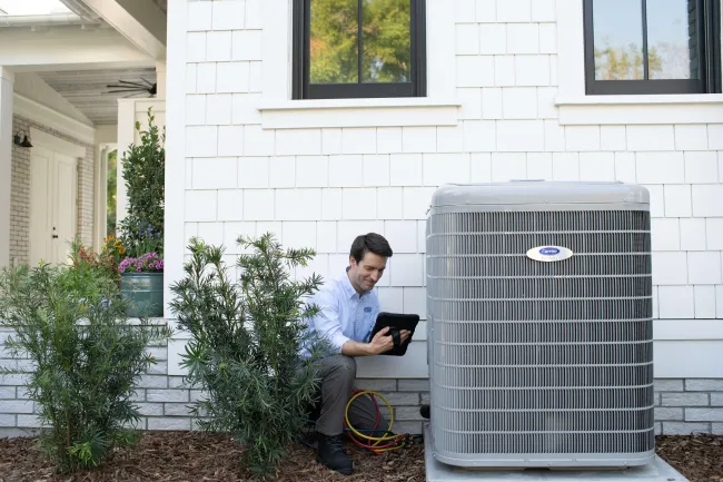 Technician inspecting a residential air conditioning unit outside a white house using a tablet device.