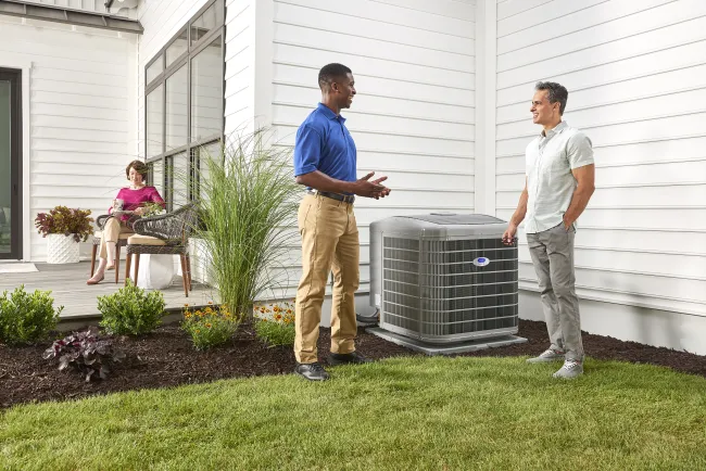 Technician discussing an outdoor air conditioning unit with homeowner outside a modern white house with garden.