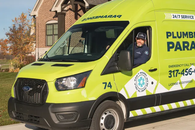 Bright green Plumbing Paramedics service van with driver smiling inside, parked in a residential driveway.