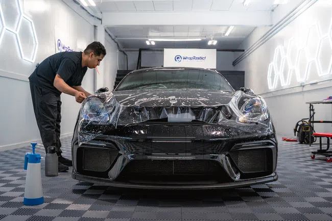 Technician applying protective film on black sports car front in a modern workshop with bright lighting.