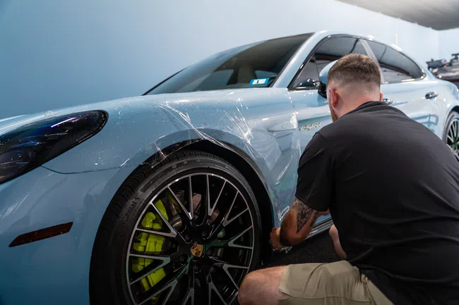 Man applies protective clear film to front side of a light blue Porsche electric car indoors.