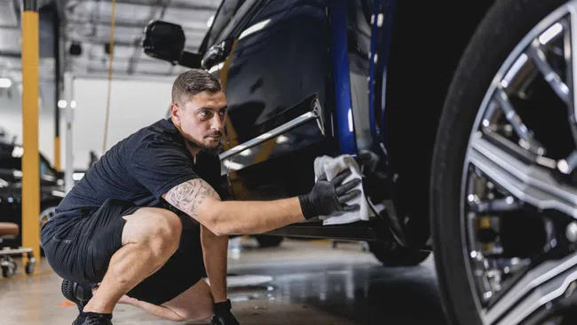 Man wearing black gloves carefully polishing a dark blue car in a well-lit garage.