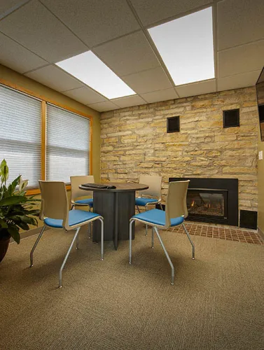 Cozy office meeting area with round table, four chairs, stone wall fireplace, and large windows with blinds.