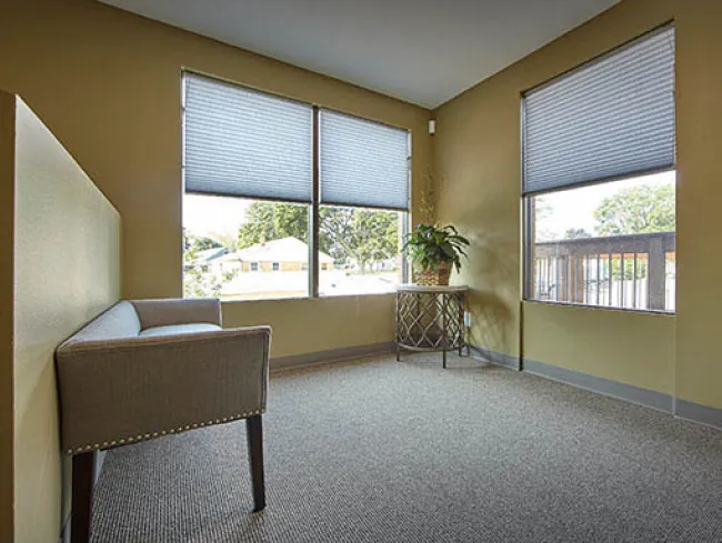 Bright room with beige walls, two windows with blinds, gray carpet, gray armchair, and decorative side table with plant