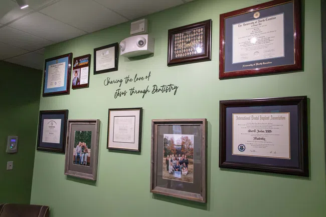 Green wall displaying framed diplomas, certificates, family photos, and a sign reading Sharing the love of Jesus through Dentistry.