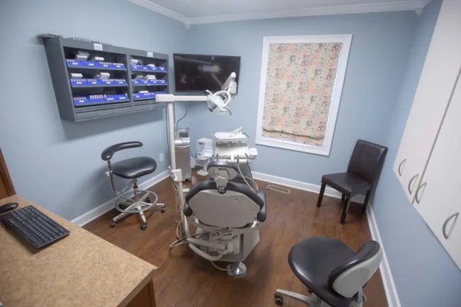 Modern dental clinic room with dental chair, equipment, monitor, stools, and storage cabinets in soft blue walls.