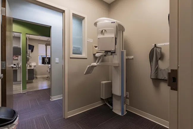 Dental X-ray machine mounted in a modern clinic with adjacent treatment rooms and equipment.