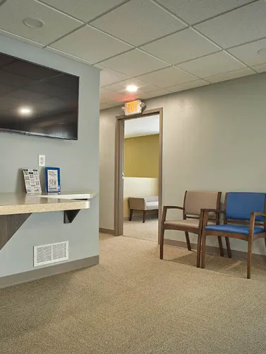 Clean waiting area with beige carpet, mixed color chairs, wall-mounted TV, and an exit door leading to another room.