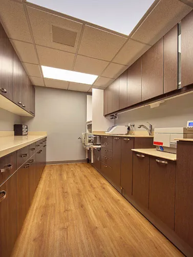 Long narrow room with wood cabinets and flooring, beige countertops, and a drop ceiling with fluorescent lighting