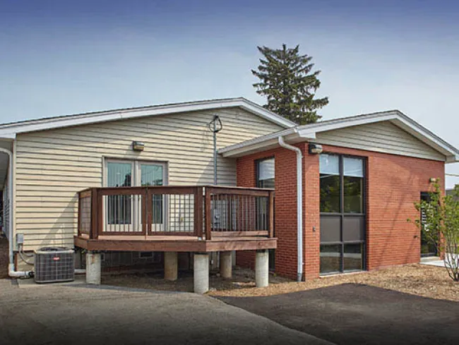 Exterior view of a modern single-story building with beige siding, brick section, and elevated deck with railing.