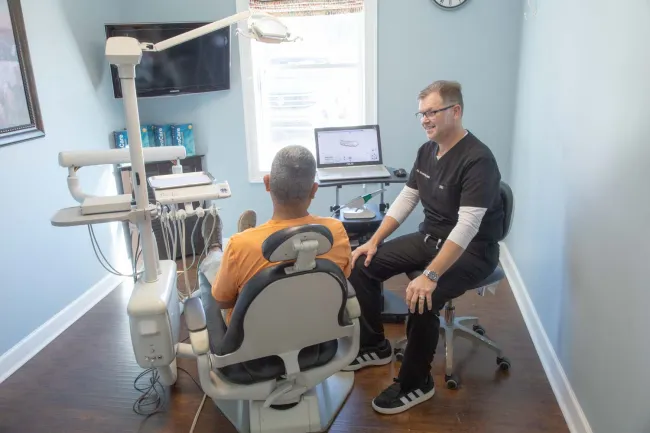 Dentist consulting a male patient in an exam room with dental equipment and digital display.