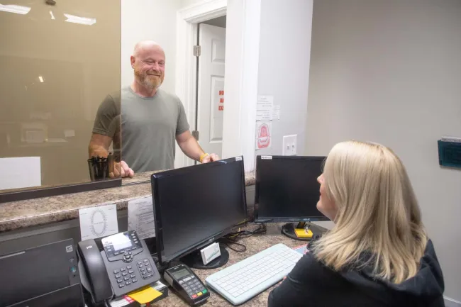 Smiling man talks to receptionist through glass window at office front desk with computer monitors and phone