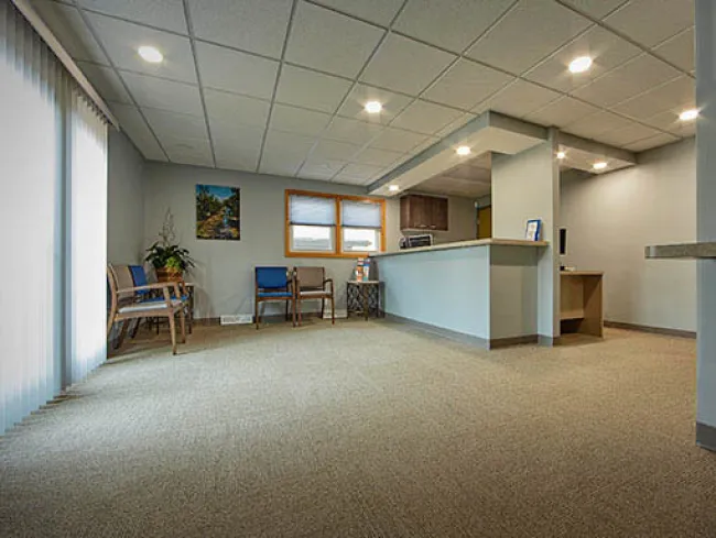 Spacious waiting room with beige carpet, blue chairs, large window, and reception counter under ceiling lights.