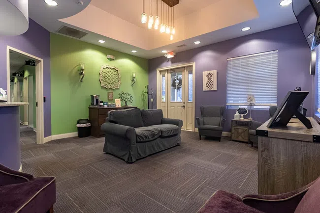 Cozy waiting room with green and purple walls, gray sofa, armchairs, decorative wall art, and soft lighting.