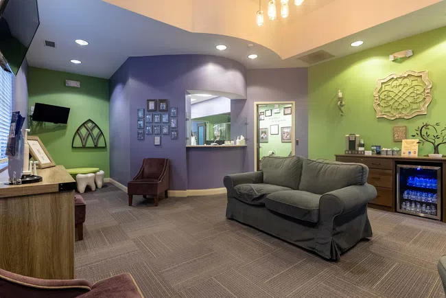 Cozy waiting room with gray sofa, purple and green walls, TV, chairs, and a refreshment station with water and coffee.