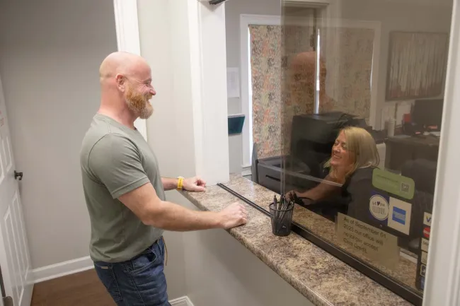 Man with beard talks to smiling woman behind protective glass at office reception desk.