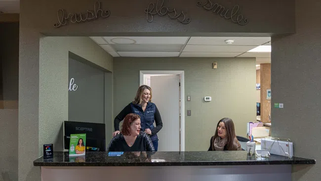 Three women smiling and interacting behind a dental office reception desk with signage saying brush floss smile