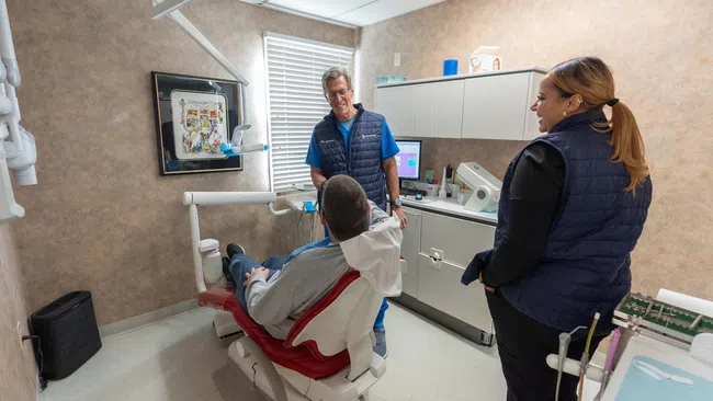 Dentist and assistant interacting with patient in a modern dental clinic with equipment and cabinetry.