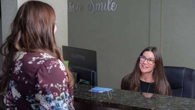 Receptionist smiling and assisting a woman at a clinic front desk with computer and sign behind.