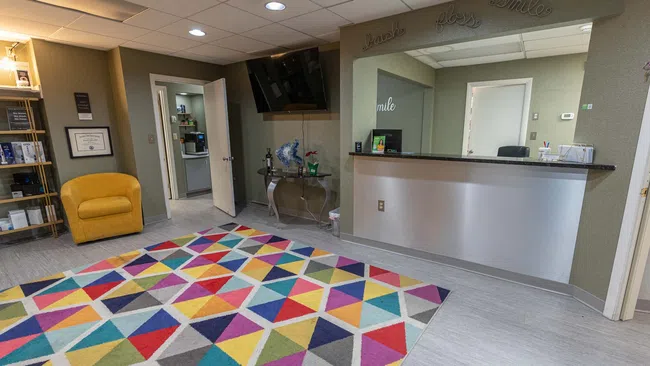 Bright dental office reception with colorful geometric rug, yellow chair, and modern desk under 'smile' decor.