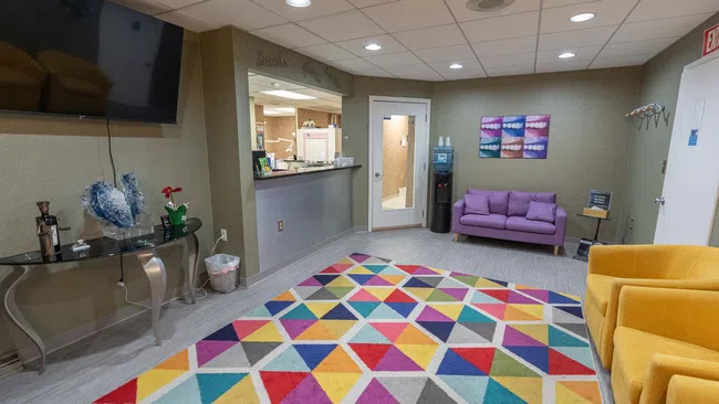 Colorful dental office waiting room with purple sofa, yellow chairs, geometric rug, and TV mounted on wall.