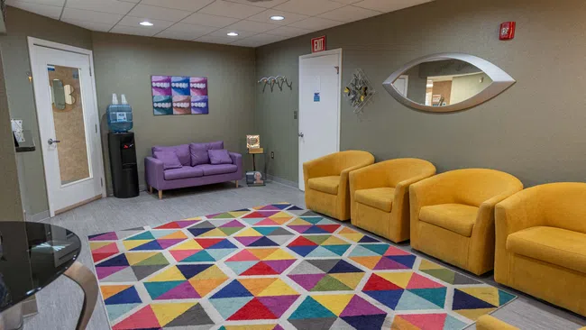 Colorful waiting room with yellow chairs, purple loveseat, geometric rug, water cooler, and decorative wall mirror.