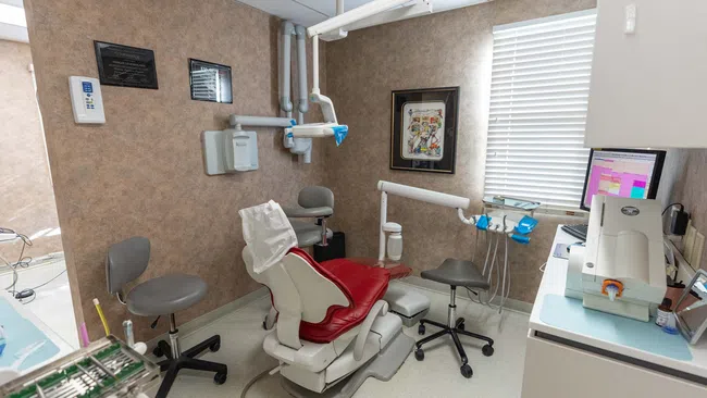 Modern dental clinic room with red patient chair, dental equipment, and a computer workstation by window