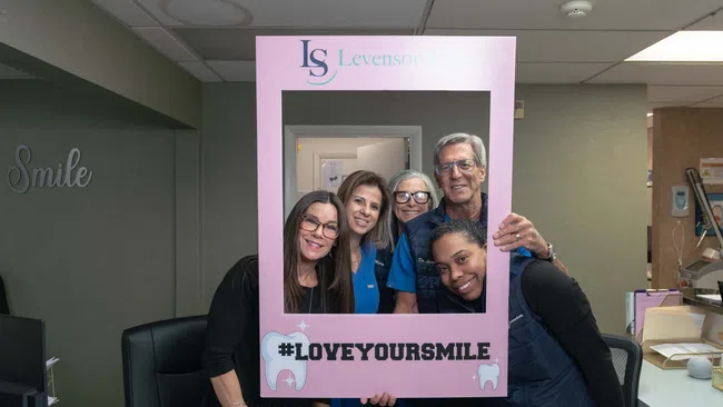 Five smiling dental staff posing behind a pink frame with #LOVEYOURSMILE at Levenson Smile clinic.
