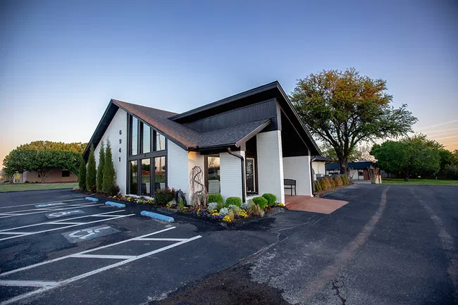 Modern white brick building with large windows, black roof, landscaped greenery, and adjacent parking lot at dusk.