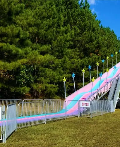 Colorful outdoor slide with pink and blue stripes beside metal railings and star decorations, green trees in background.