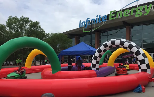 Outdoor inflatable bumper car track with colorful arches and people driving cars in front of Infinite Energy building.
