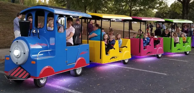 Colorful mini train with blue, yellow, pink, and green cars carrying children and adults at a park event