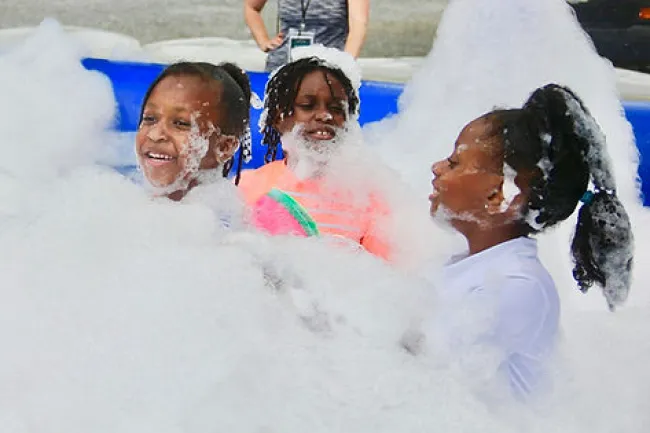 Three children playing and smiling in a large foam pit outdoors during daytime fun event.
