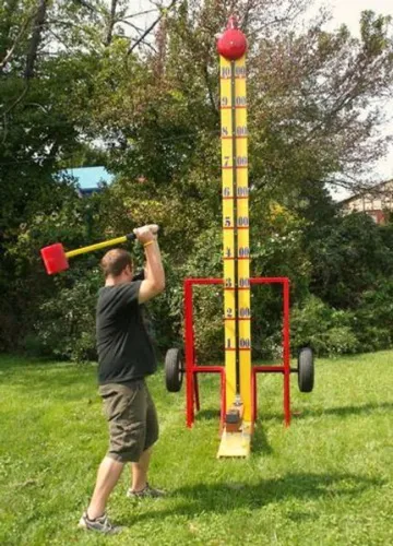 Man using a sledgehammer to hit a carnival strength tester game outdoors on green grass.