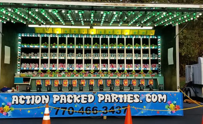 Carnival game booth with rows of stuffed animal prizes and colorful lights under green awning