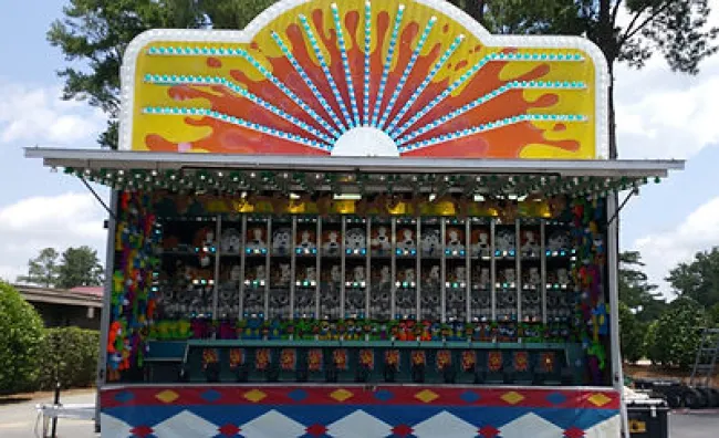 Colorful carnival game stand with prize shelves and a sunburst design on top under a blue sky