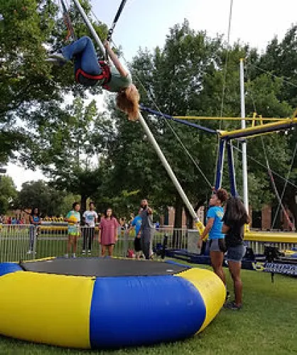 Person swinging on a bungee trampoline at an outdoor park with spectators and green trees in the background