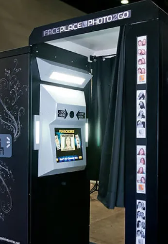 Black photo booth with illuminated screen and curtain for privacy, showing photo strip examples on the side.