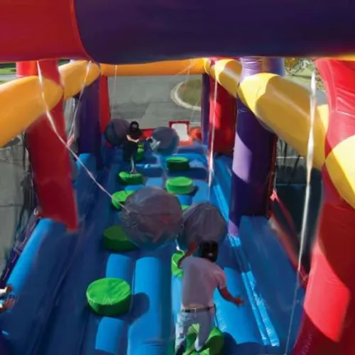 Two people navigate an inflatable obstacle course with hanging balls and green stepping pads outdoors.