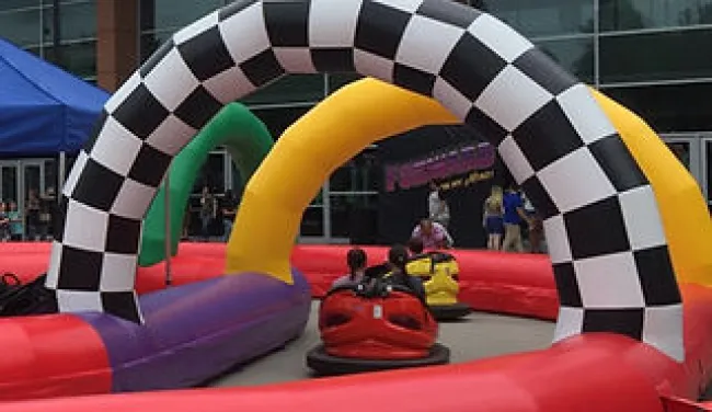 Children riding bumper cars on a colorful inflatable racetrack with checkered archways and tents nearby