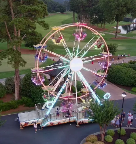 Colorful lit Ferris wheel with people at a park surrounded by trees and a golf course in the background during dusk