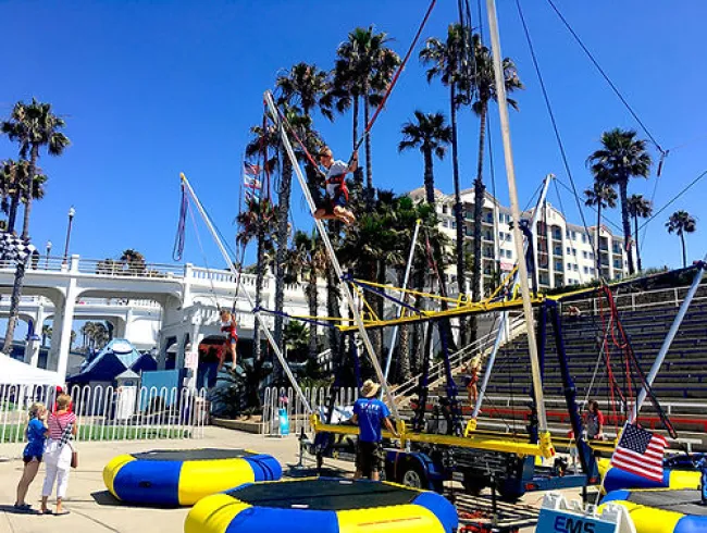 Children bouncing on trampolines and using bungee cords in a sunny park with palm trees and a white arch bridge.