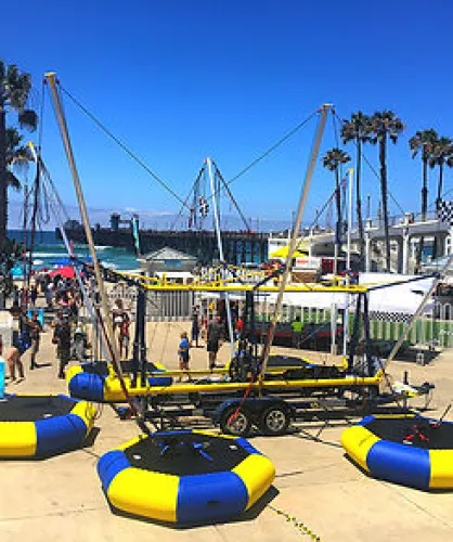 Outdoor bungee trampoline setup with yellow and blue trampolines on a sunny beach near palm trees and ocean pier.