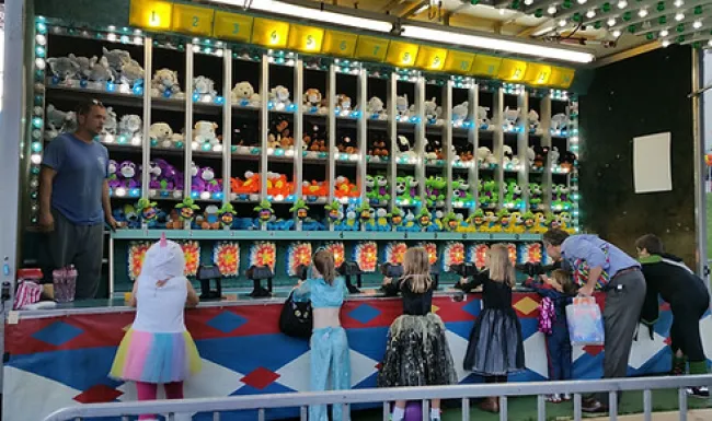Children and adults play a carnival shooting game with colorful stuffed animal prizes displayed above