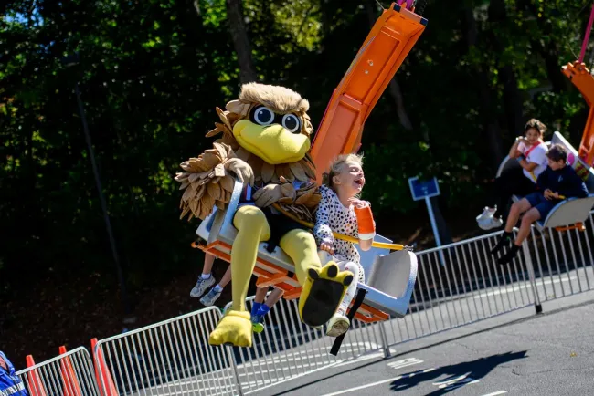 Child and mascot in bird costume enjoying a swing ride at an outdoor amusement park on a sunny day