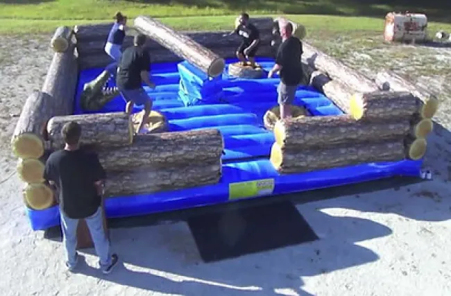 People playing on a blue inflatable arena with obstacles resembling large logs in an outdoor setting.