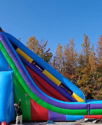 Colorful inflatable slide set up outdoors with a man standing next to it, fall trees in background under clear sky